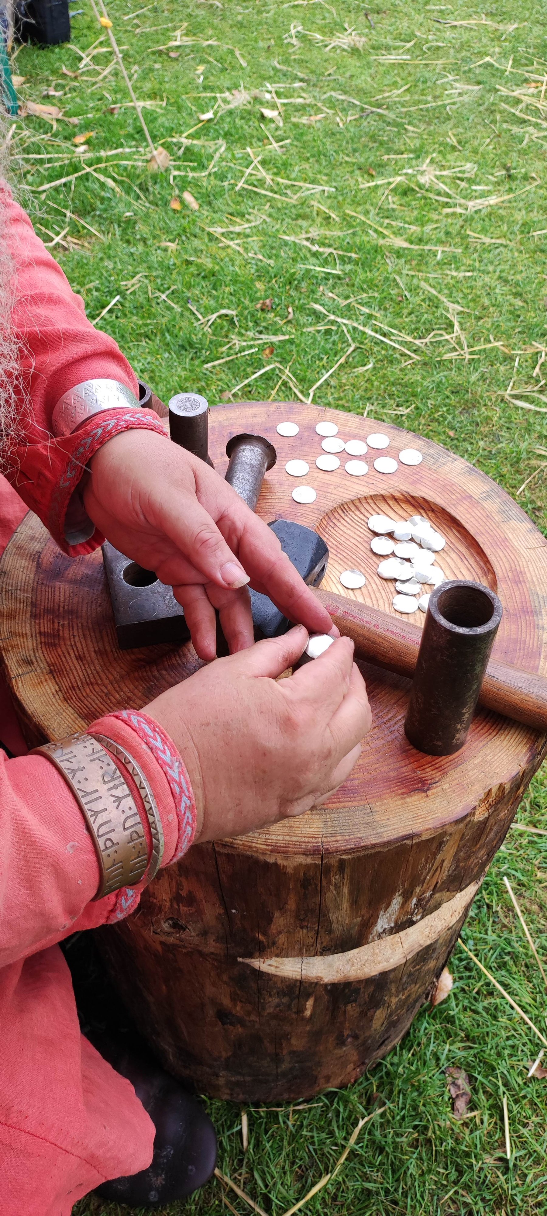A reenactor's hands striking a coin with a hammer and die on a wooden block, silver blanks scattered nearby
