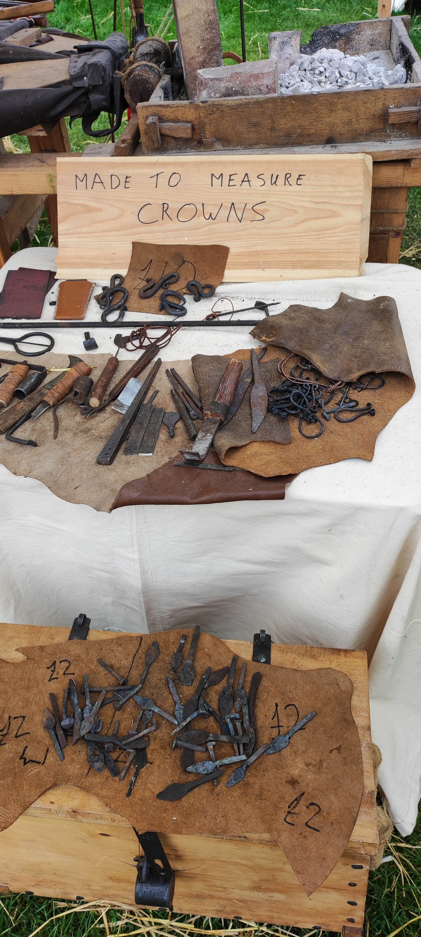 A table of hand-forged iron points and tools laid out on leather, with a "Made to Measure Crowns" sign behind them